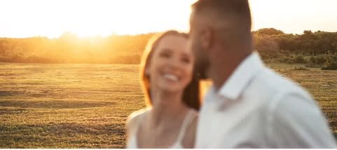 Casal sorrindo no campo, contra o sol baixo.